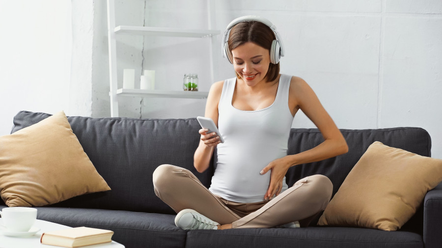 Happy Pregnant Woman Sitting On Sofa, Listening Music And Chatting On Smartphone.