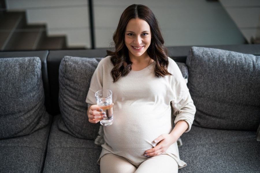 Pregnant Woman Relaxing At Home. She Is Sitting On Bed And Drinking Water.